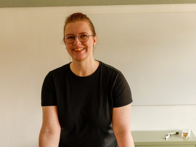 Katharina trägt eine Brille und ein schwarzes T-Shirt und steht vor einem White Board.