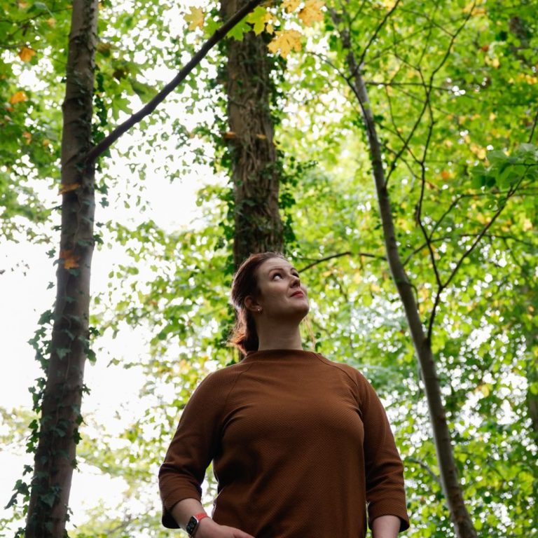 Katharina steht zwischen dünnen hohen Bäumen mit hellgrünen Blättern und schaut nach oben